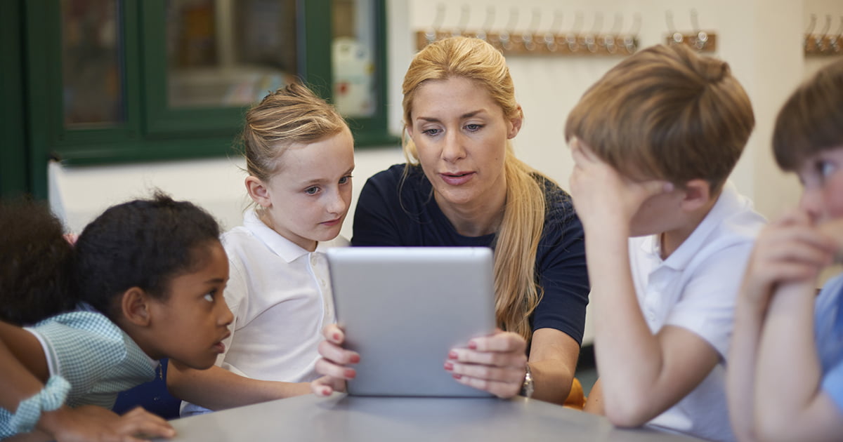 Teacher with schoolgirls and boys looking at homework on digital tablet in classroom at primary school.